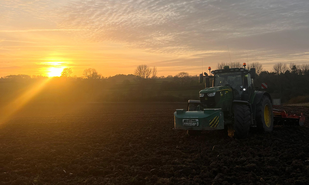 A tractor plows a dark, freshly tilled field at sunrise, with glowing orange skies and silhouetted trees in the background.