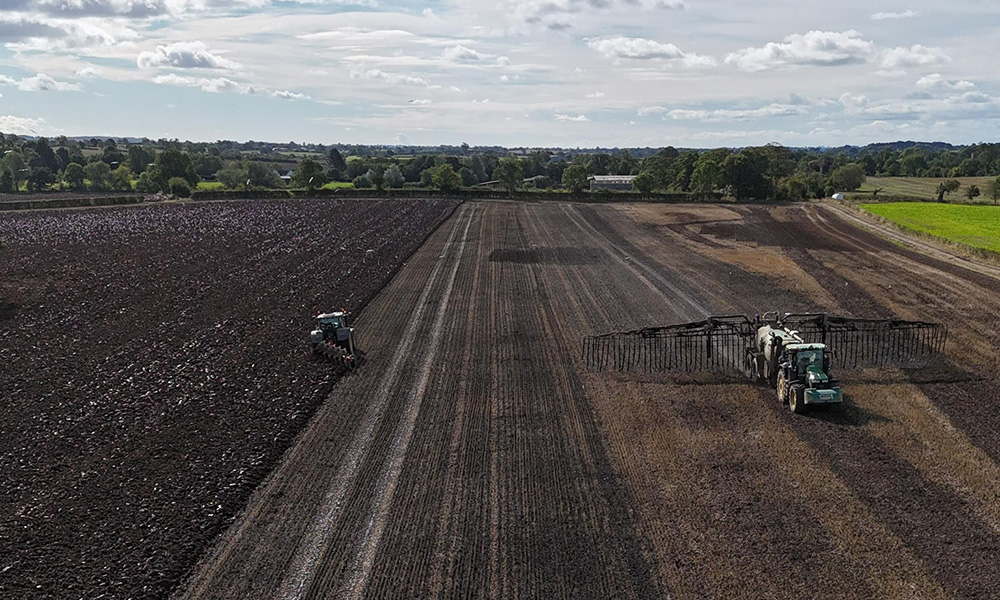 Two tractors work in a large field, one plows soil while the other uses a machine with long arms, surrounded by trees under a partly cloudy sky.