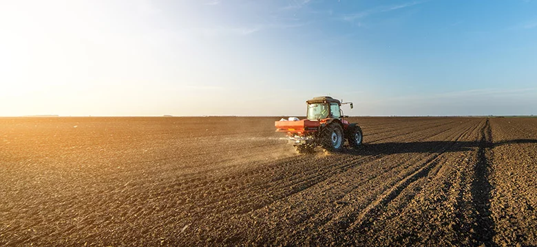 Two people shake hands in a silhouette by a cornfield at sunset, with one wearing a cap and boots, signifying a friendly exchange or agreement in an agricultural setting.