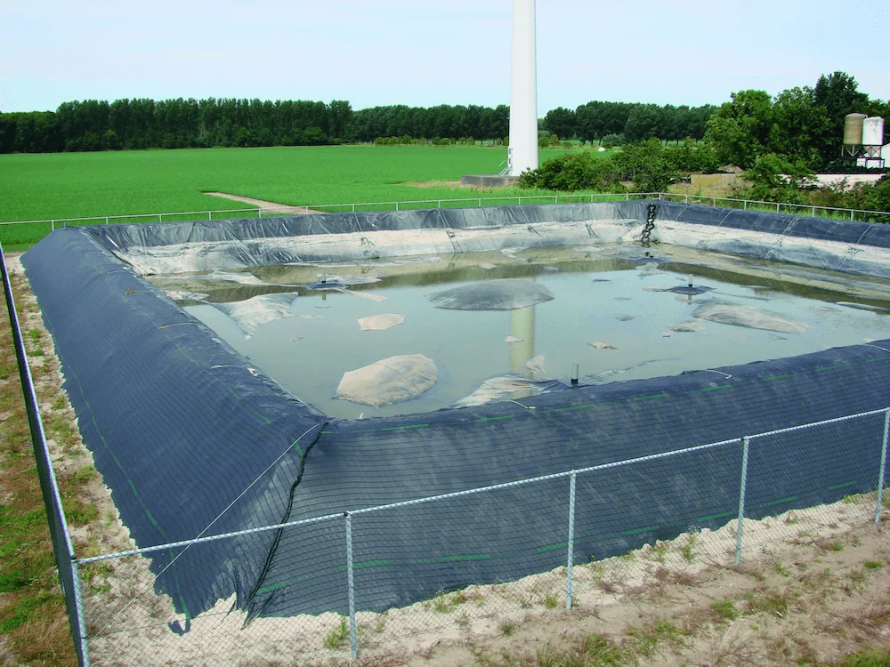 A fenced rectangular pond with a dark liner holds murky water amidst a grassy field backdrop, bordered by trees and agricultural elements in the distance, under a clear sky.